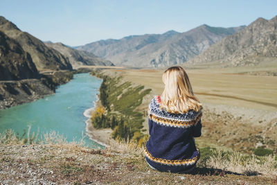 Young blonde woman in nordic sweater sitting on background of turquoise katun river, altai mountains