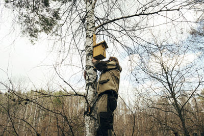 Low angle view of man adjusting birdhouse on tree trunk against cloudy sky in forest