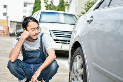 Thoughtful mechanic looking at wheel while crouching by car