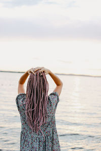 Rear view of woman with hand in hair standing at beach against sky