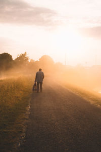 Rear view of man holding bag while walking on road against sky during sunset