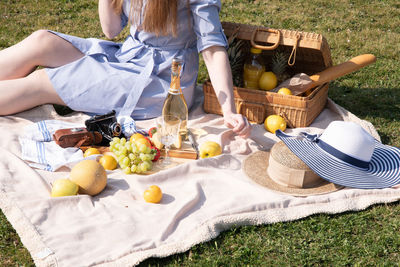 A young woman in a blue dress and straw hat is resting on a picnic with fruits