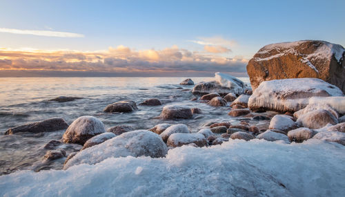 Snow covered rocks at beach against sky during sunset