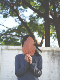 Woman covering face with dried leaf while standing against surrounding wall