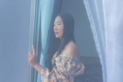 Young woman looking away while standing against window