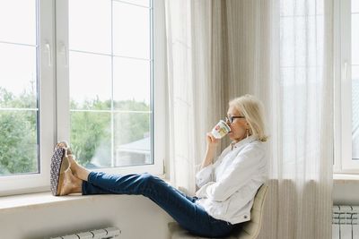 Relaxed senior woman drinking tea sitting with feet up near window at home