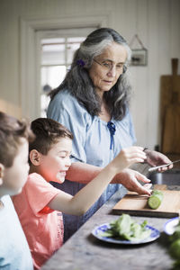 Grandmother and grandsons cutting cucumber in kitchen at home