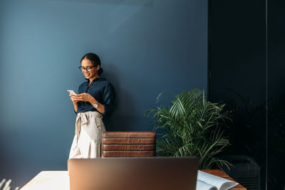 Smiling woman using phone while standing by wall in office