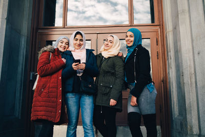 Low angle portrait of smiling multi-ethnic muslim friends standing against door