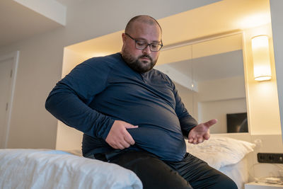 Huge man suffering from extra weight touching stomach sitting on bed in bedroom.