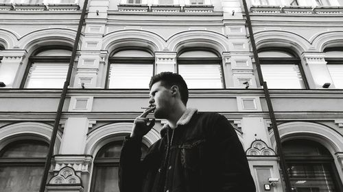 Low angle view of young man smoking cigarette while standing against building in city