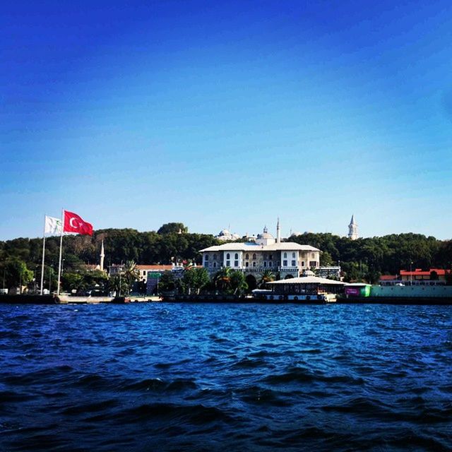 clear sky, architecture, building exterior, built structure, blue, water, copy space, waterfront, flag, patriotism, sea, day, rippled, river, identity, no people, outdoors, tree, sky, house