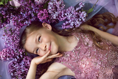 Child girl in a purple floral dress lies on the ground among lilacs on a veil in spring