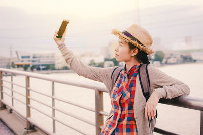 Smiling young woman taking selfie from mobile phone while standing on bridge