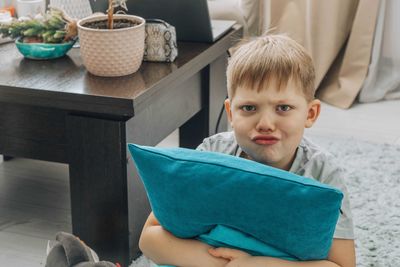 Angry boy hugs bright blue pillow in cozy home environment, looks at camera, emotion, indoors.
