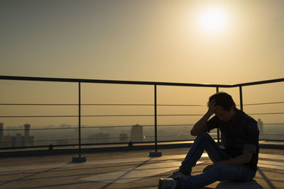 Side view of woman sitting on railing against sky during sunset