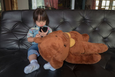 Cute girl using mobile phone with teddy bear on sofa at home