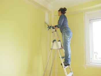 Woman on a ladder painting a yellow wall white with a paint roller