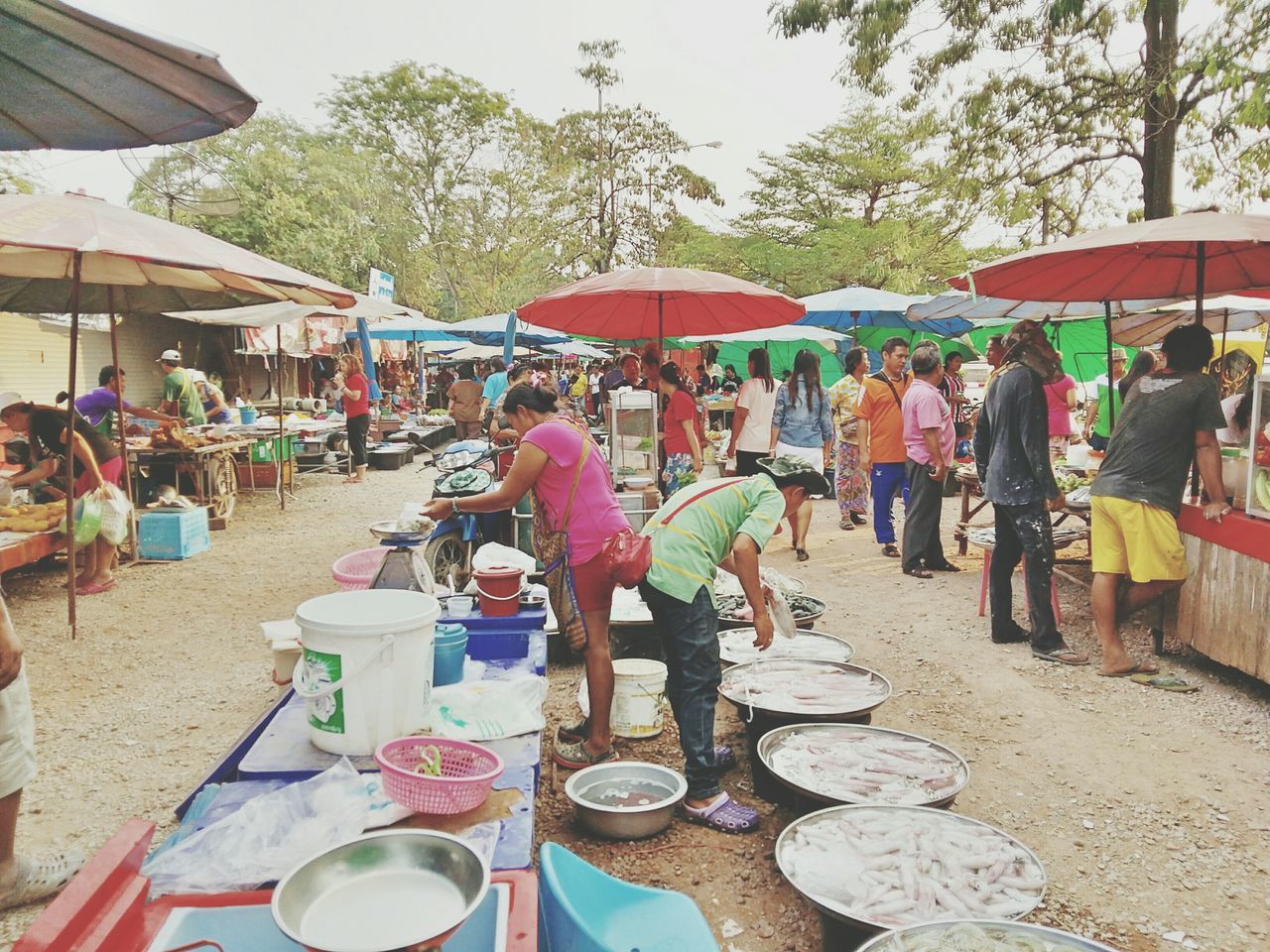 person, large group of people, men, lifestyles, tree, chair, leisure activity, market stall, day, table, large group of objects, sunlight, building exterior, outdoors, incidental people, built structure, market, sidewalk cafe, street