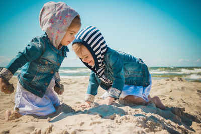 Siblings playing with sand at beach against sky