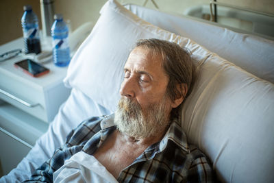 Calm aged man with beard lying under blanket on bed in hospital ward and sleeping