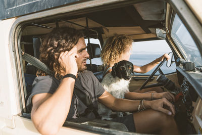 Young woman sitting by dog and man while driving car at beach