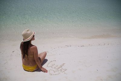 Rear view of woman in bikini sitting on shore by text at beach during sunny day