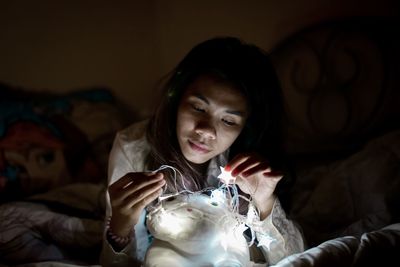 Young woman looking at stuffed toy with illuminated string lights on bed at home