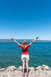 View from behind of a blonde unrecognizable tourist with her arms raised in front of lake geneva.
