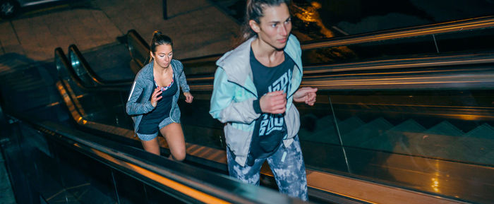 Women friends training running up escalator in city at night