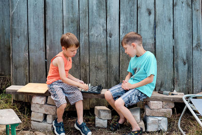 Two village boys play chess on the street, sitting on a bench, against the backdrop of an old barn