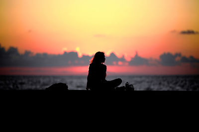 Silhouette woman sitting at beach against dramatic sky during sunset