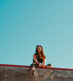Low angle portrait of smiling young woman sitting on wall against clear blue sky