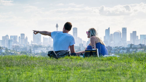 Rear view of friends sitting on grass against cityscape