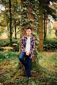 Full length portrait of smiling teenage boy standing by tree trunk in forest