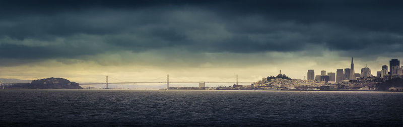 View of suspension bridge and buildings against cloudy sky