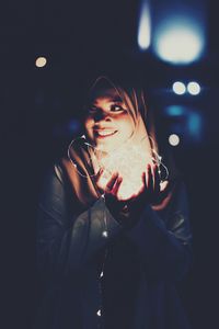 Happy young woman holding illuminated string lights at night