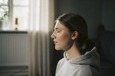 Side view of young woman looking through window at home