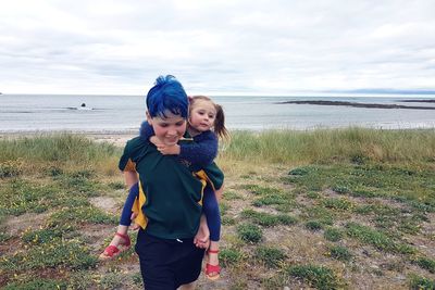 Happy brother piggybacking sister while standing on field by sea against cloudy sky