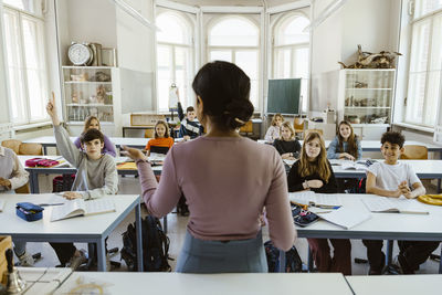 Rear view of female teacher asking students while teaching in classroom