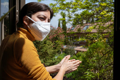 Woman wearing mask clapping while standing by railing against plants during quarantine