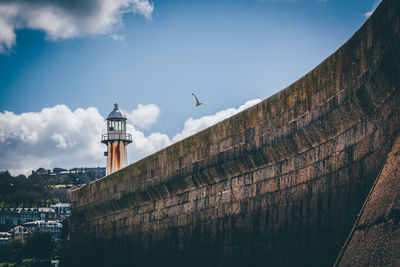 Low angle view of bird flying over wall by lighthouse against sky