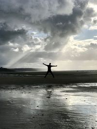 Silhouette man with arms outstretched jumping at beach against cloudy sky