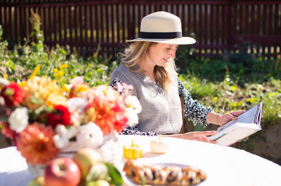 A cute girl in a hat with a brim reads a book at a table with autumn flowers on the street