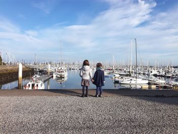 Rear view of siblings standing at harbor by moored sailboats against sky