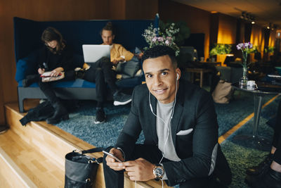 Portrait of confident businessman listening to in-ear headphones from mobile phone sitting against colleagues at office