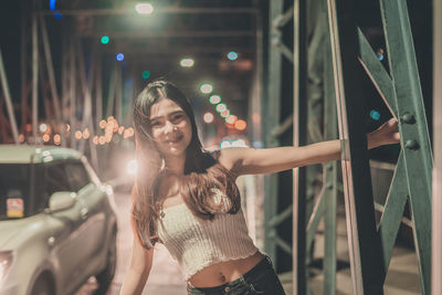 Portrait of smiling young woman standing on footbridge in city at night