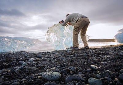 Side view of mature man photographing iceberg while standing at beach against cloudy sky