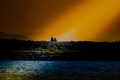 Silhouette people on rock by sea against sky during sunset