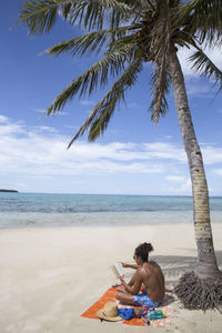 Male tourist with man bun reading under palm tree at sandy beach